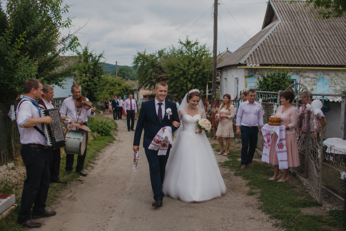 Наречений і наречена йдуть сільською вулицею під час традиційного українського весілля, поруч грають музиканти з акордеоном та скрипкою